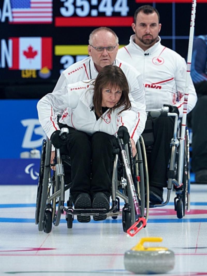 A female wheelchair curling athlete releases a stone with a stick. Four players are watching from behind her
