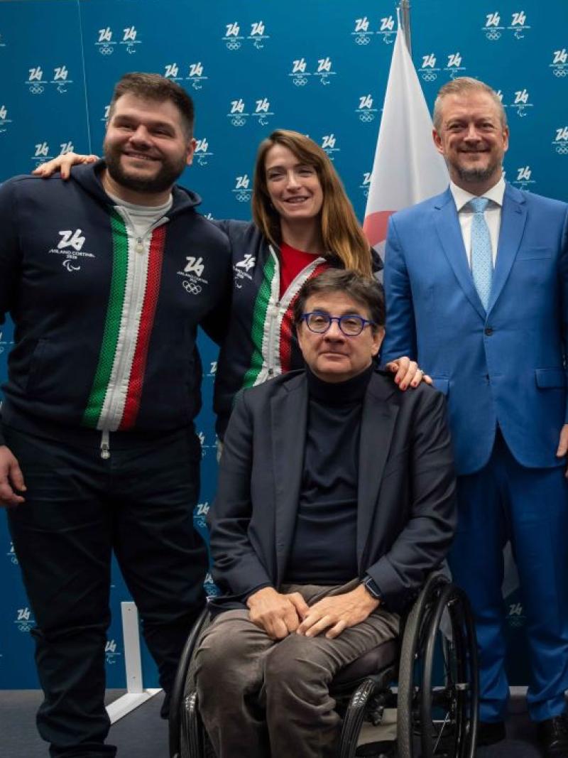 Eight people, including Paralympic athletes and officials, pose for a photograph in front of a blue banner with logos of the Milano Cortina 2026 Games