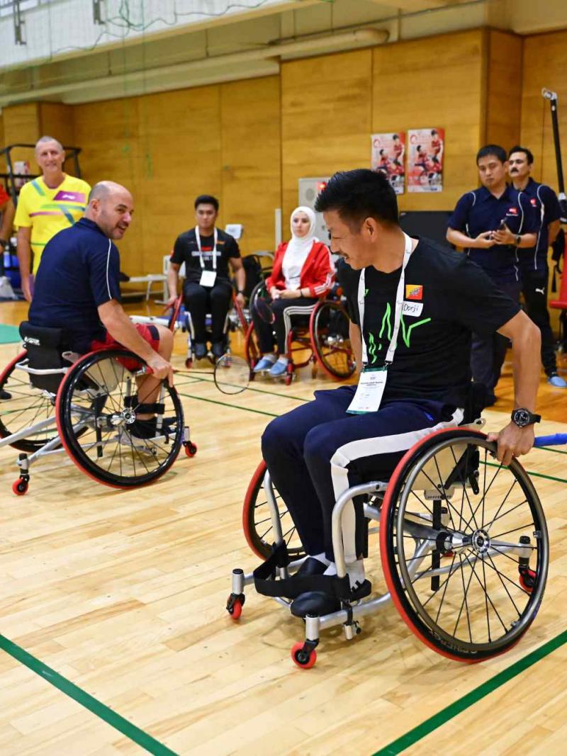 Two men holding badminton racquets are demonstrating on wheelchairs in front of 10 people