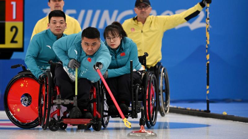 A male wheelchair curling athlete in action. He is trying to reach a curling stone with a red stick while his teammates are holding onto his wheelchair